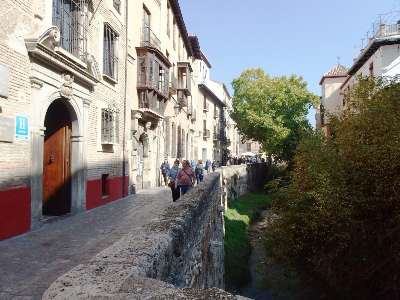 Cobbled street beside the Darro river in Granada's old town, lined with stone facades and wooden mirador balconies, with people walking along it