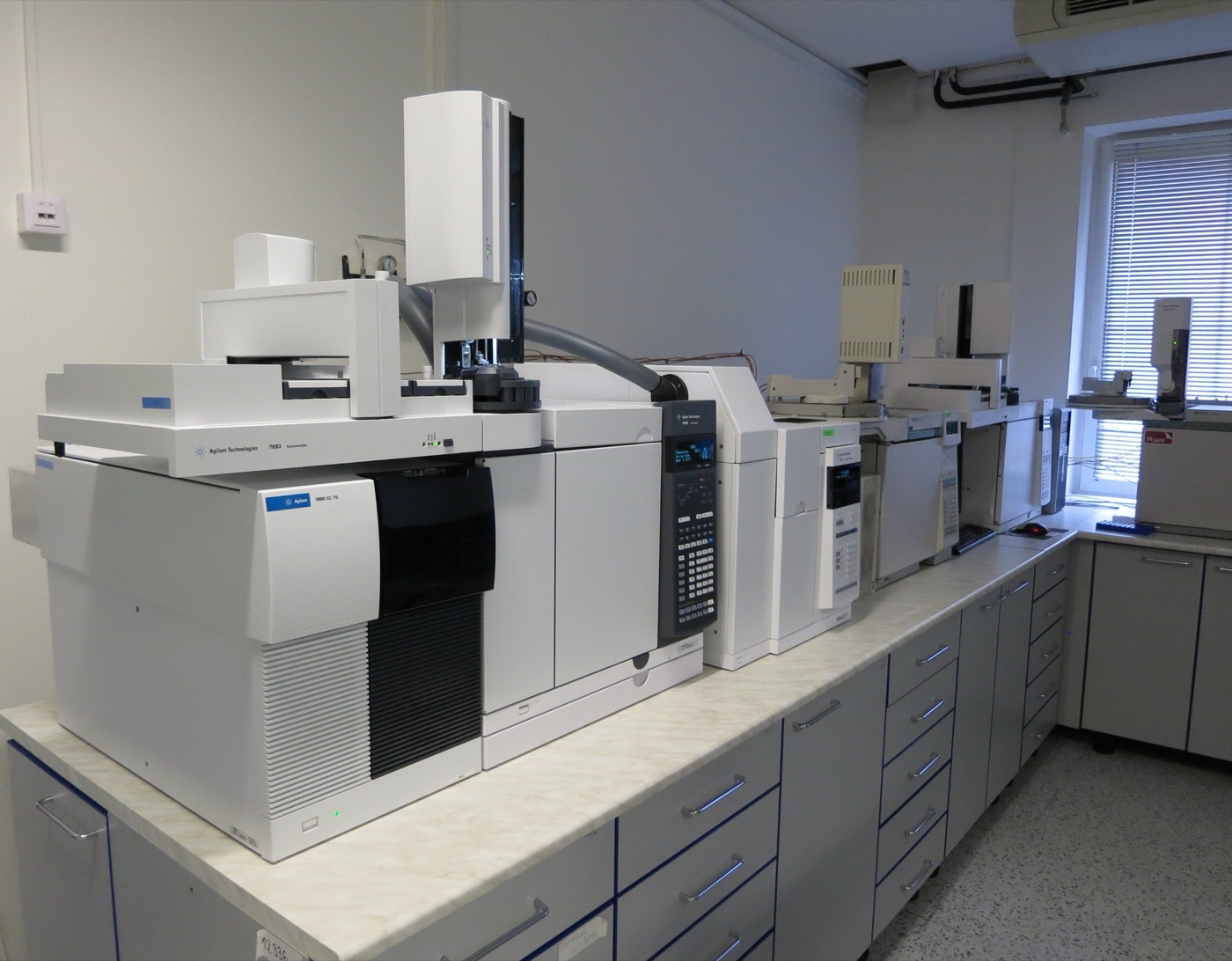A row of white Agilent gas chromatograph units with a robotic autosampler tower standing on a pale marble laboratory bench along a wall, grey drawer cabinets beneath and a blind-covered window at the far end